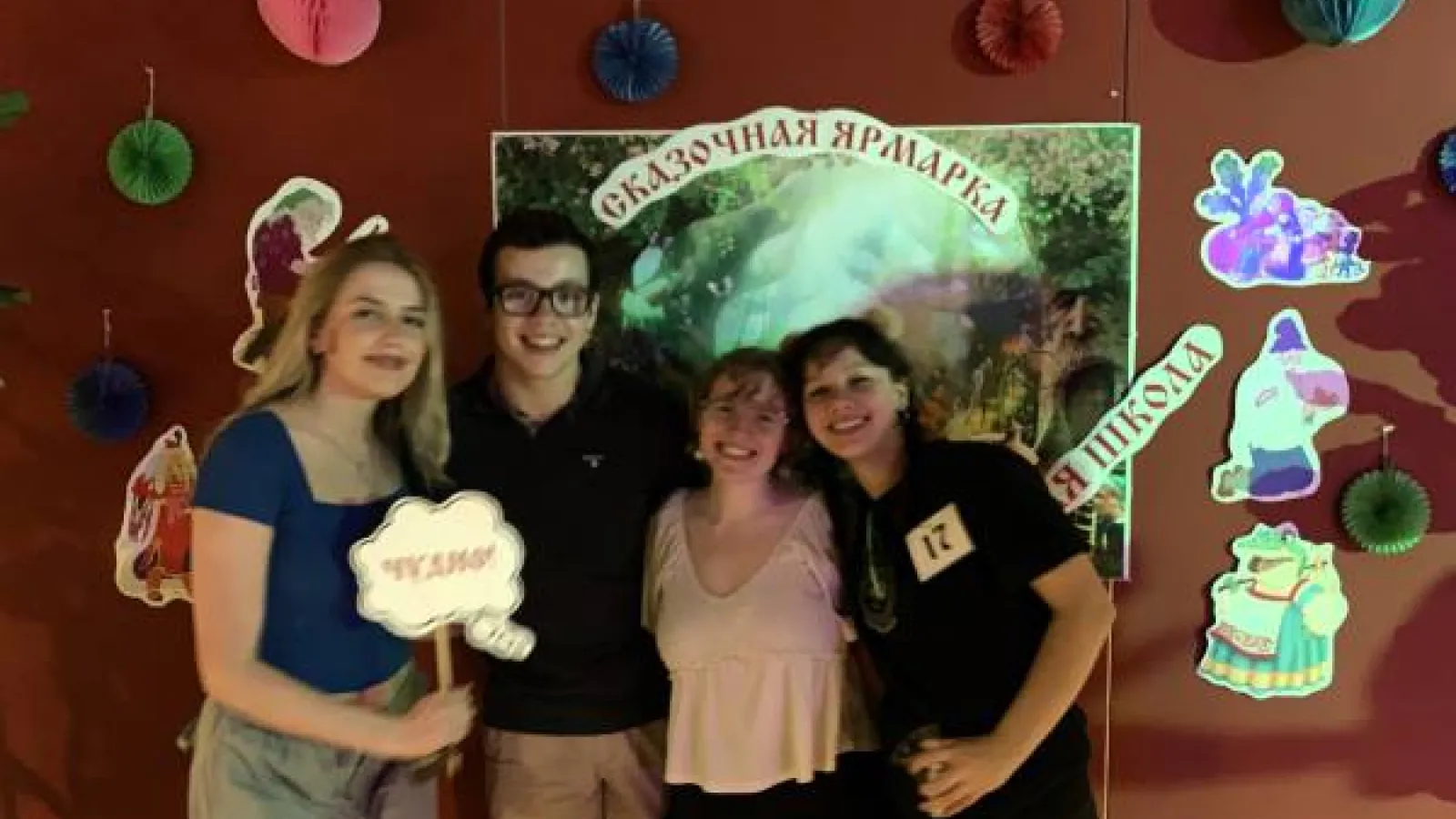 Four young people stand with their arms around each other in front of Russian-language decorations