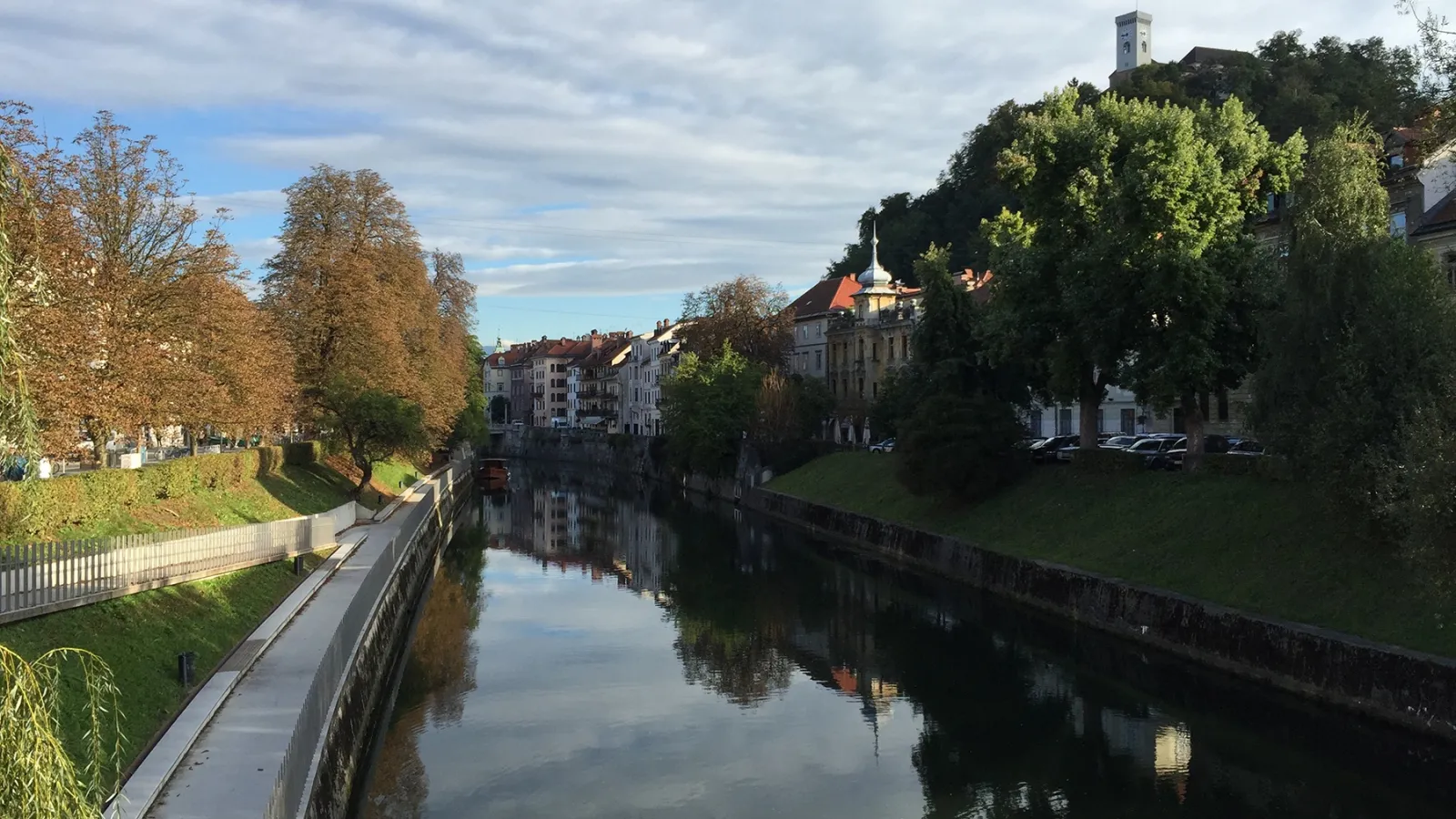 A straight river boarded by trees and grass on both sides, with buildings in the background